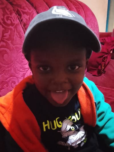The image depicts a child positioned indoors, located in what appears to be a residential setting within Kiambu Township ward, Kenya. The child, with dark skin, faces the camera directly, extending their tongue. They wear a dark grey baseball cap featuring a white, stylized bird-like emblem on the front. A black t-shirt with the yellow text "HUG YOU" and a white and grey cartoon character is visible beneath an open jacket. The jacket displays an orange interior lining and a teal-colored exterior. The background consists of deep reddish-pink upholstered furniture adorned with a prominent damask-like pattern. A segment of a lighter-colored wall is visible in the upper right, adjacent to the furniture, alongside a folded piece of dark red fabric.