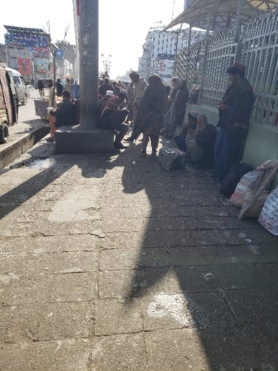 This image depicts a daytime urban scene in the city of Arghandab, Afghanistan. The foreground shows a worn, grey concrete sidewalk with visible cracks and minor debris. A dark, elongated shadow, likely from a person or object immediately out of frame, extends diagonally across the pavement. Along the right edge of the sidewalk, several white bags, some with printed text, are stacked against a green metal fence topped with a light-colored awning.

In the midground, a group of approximately 15 adult males is gathered along the sidewalk, primarily on the right side of a thick, grey concrete pole with a square base. The men are dressed in traditional attire, including long robes, shawls, and various head coverings. Several individuals are seated or squatting on the sidewalk or low concrete structures. One bearded man is squatting on the far right, observing small, light-colored rectangular objects on the pavement in front of him. Other men are standing, some leaning against the fence, while a few are seated near the base of the central pole.

To the far left, a light-colored van or truck is partially visible on the asphalt street, moving away from the viewer. The background features multi-story buildings lining the street, adorned with various billboards and signs displaying text in a local script. One partially visible sign on an upper building section reads "TB." The sky is bright and hazy, indicating strong sunlight which casts distinct shadows across the scene. The overall environment presents an active street with individuals congregating.
