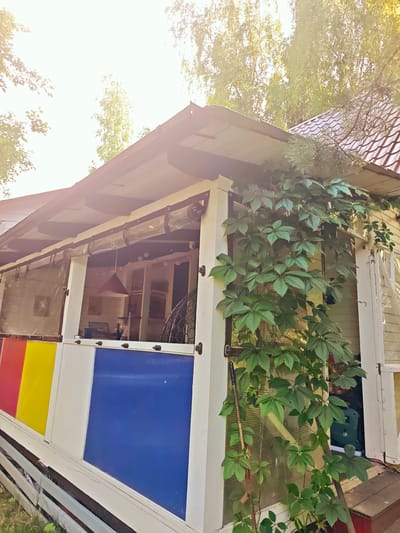 A low-angle shot captures a covered porch of a building in a rural setting, possibly a dacha in Свистуха, Russia. The building's exterior features white wooden siding and a dark-tiled roof. The porch is enclosed by transparent curtains with grommets, and a colorful section of translucent panels in red, yellow, and blue lines the lower part of the enclosure. Lush green ivy climbs the white support post on the right side of the porch, partially obscuring the transparent panels.
Through the transparent curtains, a glimpse of the interior reveals a space with various objects, including a red lampshade hanging from the ceiling, framed pictures on the walls, and what appears to be a large woven basket. A security camera is also visible near the top of the porch enclosure.
The time of day appears to be late morning or early afternoon, indicated by the bright sunlight casting a warm glow and strong shadows. The sun's rays are filtering through the leaves of trees that surround the building, creating a slightly hazy, sun-drenched atmosphere. The weather seems pleasant, with clear skies and ample sunshine. The overall scene evokes a sense of relaxed, summery outdoor living typical of a Russian dacha. There is no visible text in the image.