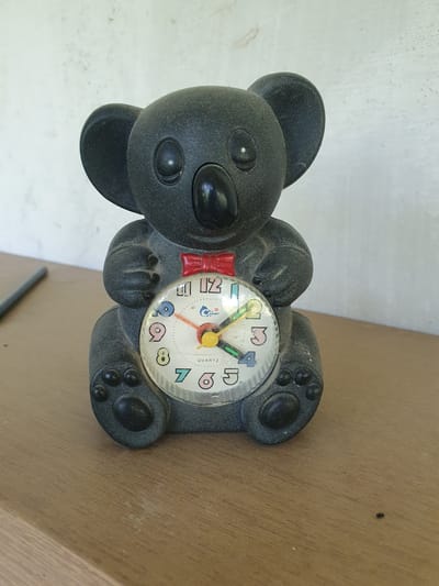 This media file features a close-up, rotated shot of a black, plastic alarm clock shaped like a teddy bear. The bear has round ears, a large black nose, and two small black eyes. It is wearing a small red bow tie. The clock face is circular and displays numbers with colorful outlines. The hour and minute hands are visible, with the hour hand pointing between 1 and 2, and the minute hand pointing to approximately the 6. The word "QUARTZ" is printed on the clock face. The bear alarm clock is resting on a light brown, textured surface, likely wood, which appears to be a table or shelf. The background is a mottled white surface, possibly a wall, with some visible dust or imperfections. The lighting suggests it is daytime, with natural light casting soft shadows. The overall impression is of a whimsical, child-oriented object placed in a simple, indoor setting. There are no people or animals in the image. The scene is static, with no apparent activity other than the presence of the object. The location context is Gomisjvari, Georgia, but this cannot be directly inferred from the visual elements.
