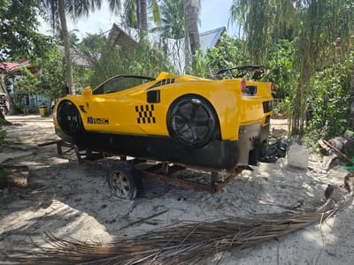 A bright yellow, novelty car body, resembling a sports car, is positioned on a rusty trailer in a sandy, outdoor area. The car body has black checkered decals on its side and what appears to be a motor attached to the rear. The trailer has one visible wheel and is resting on the sand. In the background, there are palm trees, dense green foliage, and several buildings with thatched and metallic roofs. The ground is sandy with scattered dry palm fronds. The scene suggests a tropical or beachside environment, possibly on Koh Toch Village, Cambodia, given the contextual information. There are no people clearly visible interacting with the car or trailer in the immediate vicinity. The overall impression is of a stationary, possibly decorative, object.