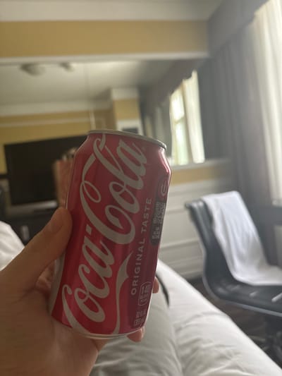 A hand holds a red Coca-Cola can. The can has white text that says "Coca-Cola" and "ORIGINAL TASTE." Below this text are the words "140 CALORIES PER CAN" and "12 FL OZ (355 mL)." The background appears to be a hotel room with a bed, a television on a stand, and a chair with a white towel draped over it. The lighting suggests it is daytime. The location is not specified in the image.