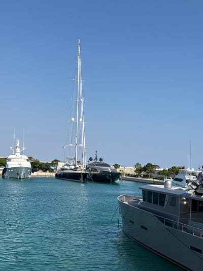 This is a landscape photograph taken in a marina in Vouliagmeni, Greece, on a clear, sunny day. The image is oriented vertically, with the bright blue sky dominating the left side and the turquoise water and several yachts filling the right. The yachts are docked along a quay lined with lush green trees and some buildings.
Several large yachts are visible. In the foreground, a sleek, dark green yacht with white detailing is moored. Behind it, a very tall sailboat with numerous white masts and rigging stands out against the sky. Further back, a large, modern, gray yacht with red trim is prominently featured, its hull reflecting in the water. Another dark-colored yacht is positioned behind the sailboat.
The water is calm with gentle ripples, suggesting a peaceful atmosphere. The sun appears to be high in the sky, indicated by the bright illumination and lack of prominent shadows, suggesting it is midday. The overall impression is one of a luxurious and serene marine setting. No people are clearly visible, and there is no visible text in the image.