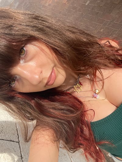 A woman lies angled with brown hair and red highlights. She wears a green top and several necklaces including one with a Star of David. Her eyes are green with light makeup. The background appears to be a brick or tile surface. The photo is taken from a low, off-center perspective.