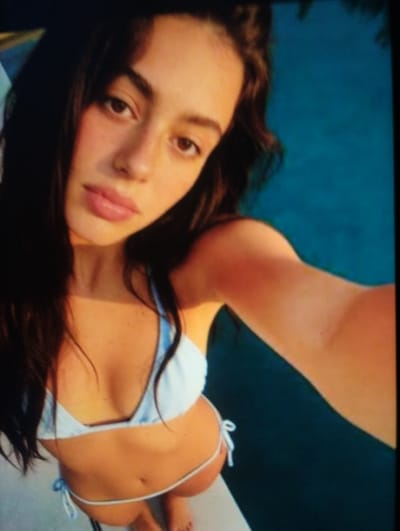 The media file shows a close-up, high-angle shot of a young woman lying down. She has long, dark hair that frames her face, which is turned towards the camera. Her eyes are open and looking directly at the viewer, and her lips are slightly parted. She is wearing a light blue bikini with white ties. Her skin is lightly tanned, and there are a few freckles visible on her face. Her left arm is raised, partially obscuring her face. The background is a blurred expanse of teal, suggesting a body of water or a pool. The lighting suggests it is daytime, and the overall impression is one of a relaxed, perhaps playful or confident moment. No text is visible in the image. The location context provided is Brashlen, Bulgaria.