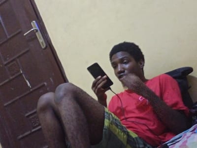 A young Black male sits on a bed, holding a black smartphone and looking at it. He is wearing a red t-shirt and plaid shorts. The background is a plain yellow wall, and to the left is a dark brown wooden door with a silver handle. The scene appears to be an indoor residential setting, possibly a bedroom, in Ukukwa, Nigeria.