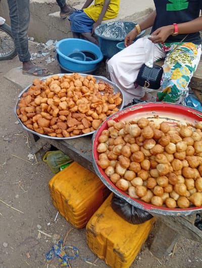 This image depicts an outdoor street or market food stall scene in Birnin Kudu, Nigeria.

In the foreground and midground, two large, round metal trays filled with fried dough items are displayed on wooden stands. The tray on the left is silver and holds numerous irregularly shaped, golden-brown fritters, consistent with *akara*. The tray on the right is red with a black rim and contains spherical, golden-brown fried dough items, consistent with *puff-puff*.

On the right side of the frame, a person, likely female, is seated. She wears a black t-shirt with a green graphic, light-colored baggy pants on her left leg, and brightly patterned floral pants on her right leg. A red watch is visible on her left wrist, and a light brown bracelet is on her right wrist. A small black shoulder bag with a gold "D" emblem rests on her lap. Her right hand holds a small clear plastic bag. Behind her, a dark plastic basin contains a white, viscous substance, likely a batter.

To the upper left, partially visible, a person, likely male, stands with dark jeans and grey sandals visible. A bicycle wheel is partially seen next to his leg. In the upper background, another person, possibly a child, is seated, wearing a yellow t-shirt and blue shorts. Additional legs and feet of another individual are partially visible behind this child. A blue plastic basin containing smaller dark items is visible in the background, near the first standing person.

The ground is unpaved dirt or sand, scattered with small pieces of debris, straw, and a knotted piece of blue and yellow patterned fabric. Two dirty, rectangular yellow plastic jerrycans are positioned on the ground in the lower right corner, beneath the food trays. The background features a simple concrete or mud-brick structure, possibly a low wall or steps.