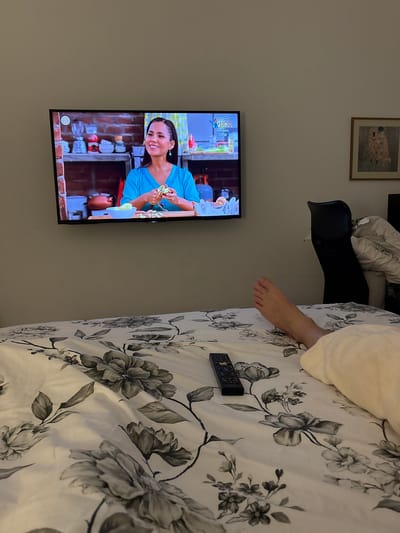 A photograph taken indoors shows a bedroom setup with a television mounted on the wall. The TV displays a woman in a blue shirt, possibly a scene from a cooking show, and is positioned on the left side of the frame. Above the television is a framed picture hanging on the wall. The right side is dominated by a bed covered with a white floral patterned duvet. A bare foot extends from under the duvet onto the bed. A black remote control is on the bed. A black head rest is visible between the wall and the bed. The location is not possible to determine from the image.