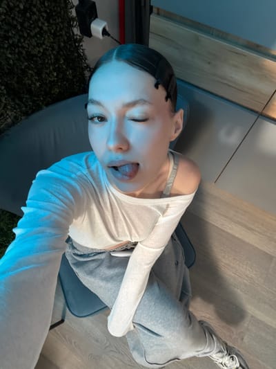A young woman is captured in a selfie taken in an indoor setting. She is wearing a white long-sleeved shirt and grey sweatpants, with a grey sports bra visible underneath her shirt. The woman has her hair pulled back and is winking, with her tongue sticking out playfully. She is sitting on a blue chair. The background shows a wooden floor, a grey wall, and a plant. A black electrical plug is also visible. The lighting suggests it is daytime or a well-lit interior. The overall mood appears casual and lighthearted. The location context is Zakharovo, Russia.