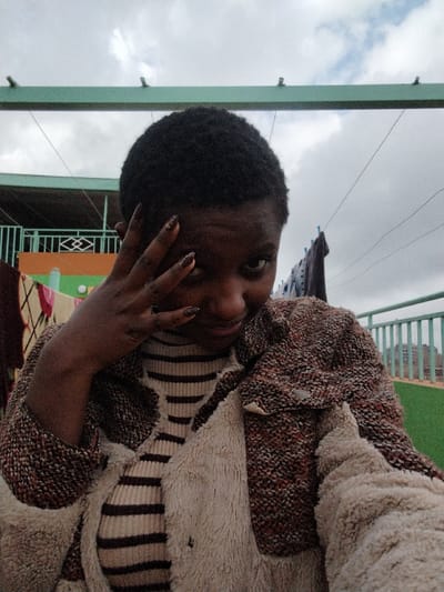 A person with short, dark hair and painted nails poses outdoors. The individual wears a striped top and a textured jacket with a fuzzy lining. The background features a cloudy sky, a green structure overhead, and a balcony railing. Laundry hangs on a line behind the person. The person's hand is near their face, partially obscuring their eye.