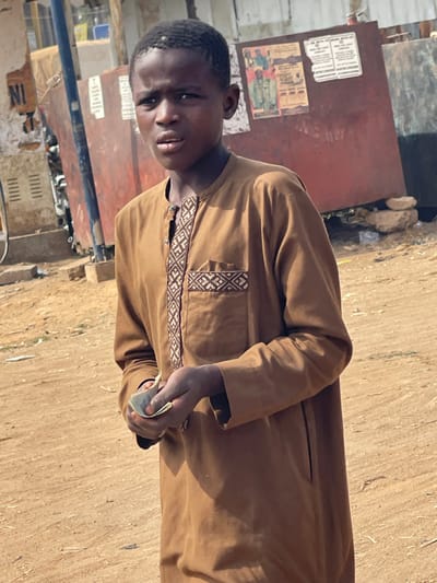A young Black male, appearing to be a pre-teen or teenager, stands on an unpaved dirt surface in Chikaji, Nigeria. He wears a light brown, long-sleeved tunic with an embroidered geometric pattern in brown and off-white along the placket and a decorative band above the left chest pocket. He holds several folded light-colored paper currency notes in his left hand at waist level. His gaze is directed forward.
In the background to the left, a blue metal pole is visible, partially obscuring a structure with a white sign displaying "NI" and "I N I". Further left, metallic elements consistent with parts of vehicles or machinery are visible. To the right, a large, reddish-brown metal panel or sheet stands vertically, tilted slightly backward. Multiple weathered paper posters or flyers are affixed to this panel; one depicts several individuals in traditional garments, while another is primarily white with black text. Small rocks are visible at the base of the metal panel. The scene is illuminated by strong natural light, casting subtle shadows.