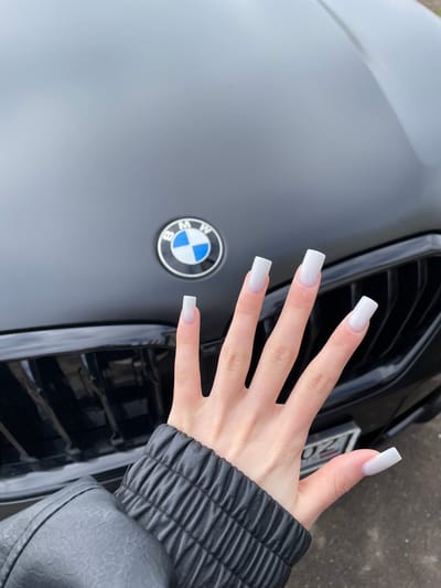 The image shows a person's hand extended over the hood of a dark-colored BMW vehicle. The hand has long, square-shaped, white nails with a subtle shimmer or glitter effect. A portion of a dark, quilted jacket sleeve is visible near the wrist. The BMW's hood, featuring the circular BMW logo, and its black grille are prominent. The license plate is partially visible. The location is given as Karmaskaly, Russia, but the specific details suggesting this location are not discernible from the image itself.
