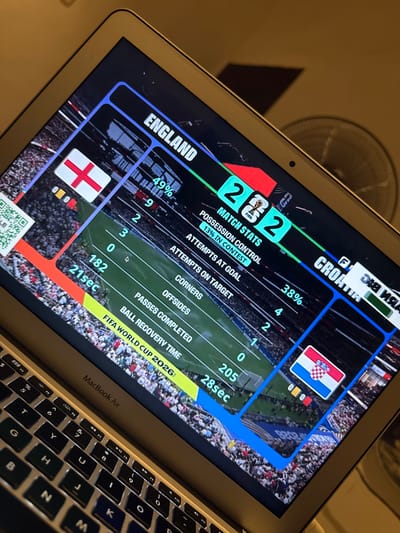 The image shows a close-up of a MacBook Air laptop screen displaying a football (soccer) match simulation. The screen shows the "FIFA World Cup 2026" graphic. The match appears to be between England and Croatia. England has the flag shown and the score is 2-2. Match stats for England are displayed: 49% possession, 9 attempts, 2 shots on target, 3 corners, 0 offsides, 182 passes completed, and a ball recovery time of 21 seconds. Croatia's flag is also visible, with a score of 2-2, and their possession is 38%, with 4 attempts, 2 shots on target, 1 corner, 0 offsides, 205 passes completed, and a ball recovery time of 28 seconds. The background of the screen shows a stadium filled with spectators. The laptop keyboard is partially visible in the foreground. The image is taken at an angle, and the lighting suggests an indoor setting, possibly in the city of Kufena, Nigeria, although this location is not explicitly shown in the image.