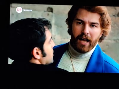 The image shows two men in a close interaction. One man has dark hair and is wearing a black shirt, with only the side of his face and ear visible. The other man, facing the viewer, has reddish-brown hair and a beard. He is wearing a white turtleneck sweater underneath a bright blue blazer. The scene is likely from a television show, indicated by the "DF премьера" logo in the upper left corner of the image. The background is blurred, but there appears to be a concrete wall. Based on the location in the city of Zverino, Bulgaria, the scene could be a segment from a television series or movie. The men seem to be in conversation, with the man in the blue blazer possibly speaking.