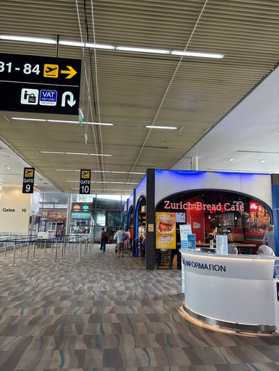 The image depicts an interior scene at an airport, likely within Mai Khao, Thailand, given the context provided. The environment features a modern, tiled ceiling with integrated lighting. A prominent black sign indicates "31-84" with directional arrows and symbols for a plane and VAT refund. Below this, a sign for "GATE 9" is visible, followed by another sign for "GATE 10." The floor is covered in a patterned carpet with abstract blue and gray wavy lines.
To the right, there is a "Zurich Bread Café" with illuminated signage. A yellow poster advertising "Healthy & Yummy!" with a sandwich is displayed. Next to the café, a white, curved information desk with the word "INFORMATION" in blue letters is positioned. Two individuals, appearing to be airport staff, are visible behind the desk, with one wearing a headscarf. Several travelers are walking through the area, with some queuing at what appear to be security or boarding gates, marked by blue velvet ropes and stanchions. In the background, elements of other shops or concessions, including "BURGER KING" and "CAMDEN FOODS," are partially visible. The overall scene conveys a busy, functional airport terminal.