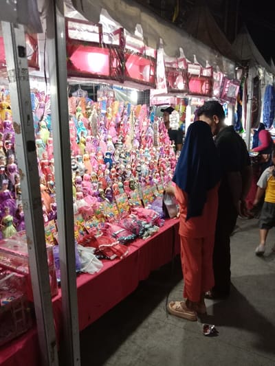 The image shows a street market stall in Kaliwungu, Indonesia (latitude: -6.962, longitude: 110.252). The stall is selling dolls and other toys. There are many rows of dolls on display, all dressed in colorful clothing. A woman wearing a blue head scarf and orange pants is looking at the dolls. A man in a black shirt and a blue head scarf is standing behind her. A second man in a black shirt is standing behind the first man, with his back to the viewer. They appear to be looking at the dolls. There are several other people in the background of the image. The stall is covered by a white canopy, and there are lights overhead. The ground is a concrete surface.