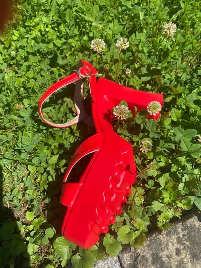 A bright red, platform sandal is lying on a bed of green clover and grass, with a few small white clover flowers interspersed. The sandal has a thick, chunky sole with a raised, textured pattern on the toe area. A single ankle strap, also red, is visible, fastened with a silver buckle. Part of the sandal's heel is elevated, suggesting it might be a wedge or platform heel. In the upper right corner of the image, a rough, greyish-brown concrete or stone surface is visible, possibly a pathway or wall. The lighting indicates it is daytime, with strong sunlight casting shadows. The scene appears to be outdoors, in a natural or garden setting. No people or text are visible in the image.