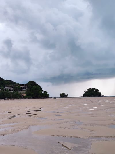 This image captures a coastal scene in Sakhu, Thailand, characterized by a wide, sandy beach at low tide. The sand is wet and rippled, with numerous shallow pools reflecting the overcast sky. In the distance, two small, tree-covered islands are visible. The larger island on the right has a dense canopy of trees, while the smaller one on the left appears to have only a few trees.
On the left side of the image, a lush, green hillside overlooks the beach. Abandoned, multi-story buildings with exposed concrete structures are nestled into the vegetation on the slope, suggesting a former development or construction project. The buildings are overgrown with trees and vines, blending into the natural landscape. A small boat is moored on the shoreline near the hillside.
Two figures are walking on the beach in the middle ground, appearing as small silhouettes against the sand and water. The sky is heavily overcast with dramatic, dark clouds that suggest an impending storm or recent rainfall. The overall atmosphere is moody and serene, with a sense of nature reclaiming man-made structures.