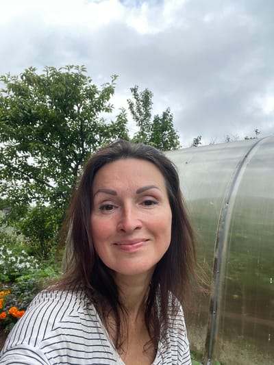 A selfie of a woman taken outdoors in Дресвяново, Russia. The woman, with brown hair and fair skin, is smiling. She is wearing a white shirt with black stripes. Behind her, to the left, is a green tree. To the right is a greenhouse. The sky is overcast.