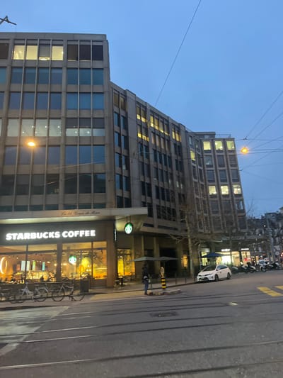 The image captures an urban street scene in Geneva, Switzerland, during dusk or early evening. A prominent multi-story building, appearing modern with a concrete and glass facade, dominates the left side of the frame. Many windows of this building are illuminated with warm yellow light. The building features an interesting architectural design where its upper section appears to recede or step back.
On the ground floor of this building, a brightly lit Starbucks Coffee shop is visible, with its large glass windows revealing the interior. Several bicycles are parked in a rack directly in front of the Starbucks entrance. Along the right side of the image is a street or road, where a white car is parked, and several scooters or motorcycles are also visible further down the street. At least two individuals can be seen on the sidewalk near the Starbucks, one appearing to walk, the other potentially standing. Bare tree branches suggest the season might be winter or early spring. Overhead wires, possibly for public transport such as trolleybuses, crisscross against the twilight blue sky.
Visible text on the Starbucks facade clearly reads "STARBUCKS COFFEE." Below the Starbucks logo, the address "Le 11, Cours de Rive" is also partially visible on the building. The overall scene depicts a typical evening in a bustling city.
