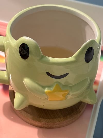 A light green ceramic mug, designed to resemble a frog with black eyes, a black smile, and a yellow five-pointed star on its chest, holds a translucent light amber liquid. The mug rests on a circular wooden coaster with visible concentric grain patterns. Both the mug and coaster are positioned on a smooth pink surface. To the right, a portion of a white object with a blue edge is visible. The background exhibits indistinct, soft-focus colored areas, including hues of pink, green, and blue. The scene is located in Voluyak, Bulgaria.