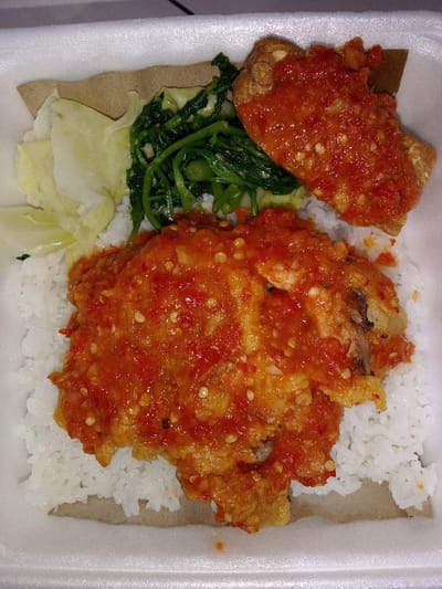 The image displays a traditional Indonesian meal, likely a takeout or street food dish, presented in a white styrofoam container. The main component is a large serving of fluffy white rice. Topping the rice are two pieces of what appears to be fried chicken or fish, generously covered in a vibrant, chunky red chili sauce, commonly known as 'sambal'. The sauce is rich in texture with visible chili flakes and seeds, indicating a spicy flavor profile.

Accompanying the main protein are two types of vegetables. On the left side of the container, there are several pieces of light green to white cabbage, which appear to be steamed or blanched. Adjacent to the cabbage and above the rice, a portion of dark green leafy vegetables, possibly water spinach (kangkung), is visible. A piece of brown paper or similar natural lining, potentially a banana leaf, is placed under some of the food, adding an earthy tone to the presentation. The dish is characterized by its contrasting colors: the pristine white of the rice, the intense red of the chili sauce, and the various greens of the vegetables. No text is visible in the image. The meal reflects typical Indonesian culinary elements, consistent with the given location context of Situbondo, Indonesia.