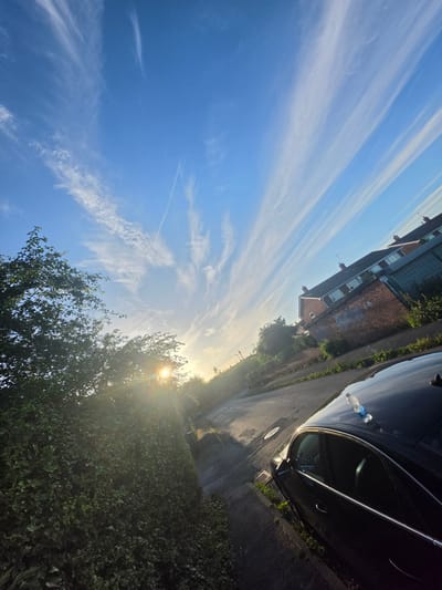 A sunlit, low-angle outdoor shot captures a suburban street scene in Shotton, United Kingdom. The sky, a brilliant blue, is streaked with wispy white clouds that fan out from the horizon. The sun, a bright orb with visible lens flare, is low in the sky, casting long shadows and illuminating the scene with a warm glow.
On the left side of the image, a dense, dark green hedge lines the street. Behind the hedge, leafy trees are visible, their branches reaching towards the sky. The sunlight filters through the leaves, creating dappled patterns.
A black car is parked on the right side of the road, angled towards the viewer. A plastic water bottle is placed on its roof. The car's windows are tinted, and the interior is mostly obscured. The car is positioned on the asphalt roadway, which appears wet in places, reflecting the sky and the sunlight.
The background features houses with red brick walls and dark tiled roofs, typical of residential architecture in the area. Some of the houses have windows, and various architectural details, such as chimneys and gutters, are discernible. Greenery, including overgrown grass and bushes, can be seen around the houses and along the side of the road. A few manhole covers are visible on the pavement. The overall impression is one of a quiet, residential street bathed in the soft light of a morning or late afternoon sun.