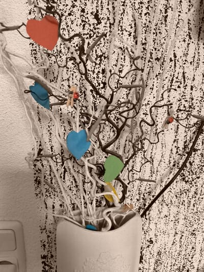 The image shows a decorative arrangement in a vase. The arrangement consists of white branches and dark, curly branches, adorned with several colorful paper hearts (red, blue, and green) and some small decorative ladybugs attached with clothespins. The background is a textured wall with a dark, speckled pattern. The overall impression is a rustic and whimsical decorative piece. There is no indication of the location being Dillingen an der Donau, Germany.