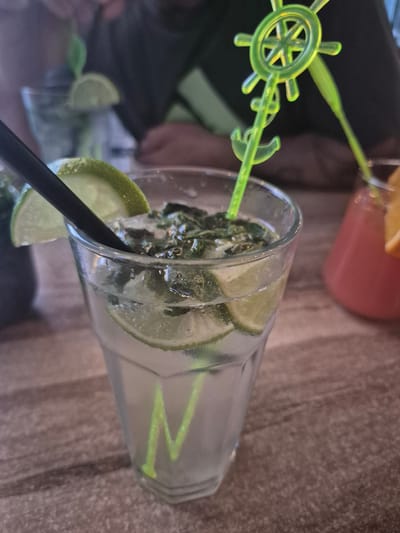 A close-up, canted shot shows a cocktail with lime wedges and a straw in a glass. The drink is on a surface with a textured, slightly blurred background. A second drink in a pinkish hue is visible in the background with a green plastic decoration of a ship's wheel and anchor. There is no visible evidence to indicate a specific location in Pavlikeni, Bulgaria.