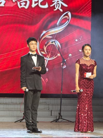 This image captures two individuals, a woman and a man, standing on an indoor stage, likely hosting or presenting at a formal event in Guta, China.
The background is dominated by a large, illuminated red screen or backdrop. It features a prominent golden treble clef and scattered musical notes, hinting at a musical or performance-related occasion. On the left side of the screen, stylized white Chinese characters are visible, reading "梦想飞" (mèngxiǎng fēi), which translates to "Dream to Fly" or "Flying Dreams."
The woman, positioned on the right side of the frame, wears an elegant, floor-length reddish-maroon gown adorned with sequins in a zigzag pattern and intricate beading across the shoulders. She has dark hair, delicate drop earrings, and holds a microphone in her right hand and a small white card in her left, suggesting she is speaking or reading from a script. She maintains a poised, gentle smile.
Beside her, the man is dressed formally in a black tuxedo with a white bow tie and glasses. He holds a microphone in his left hand and a small reddish card in his right, mirroring the woman's activity. He also presents a composed, slight smile.
Both individuals appear professional and engaged in their roles, suggesting they are hosts or key participants in an event, possibly a concert, award ceremony, or cultural performance. The overall setting is brightly lit, typical of an evening stage event. The stage floor is partially visible on the far right, appearing to be a light-colored surface.