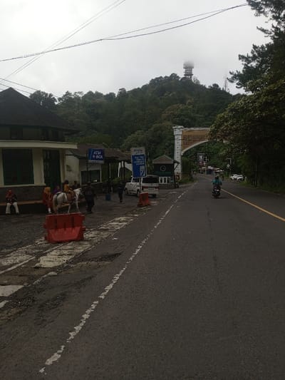A road leads towards a mountainous area, featuring an archway structure. A communications tower is visible atop the hill, partially obscured by clouds. A building sits on the left, near parked vehicles and people. A horse stands near orange barriers. A motorbike travels along the right side of the road, alongside a yellow line. ATM and Electric Vehicle Charging Station signs are also present.