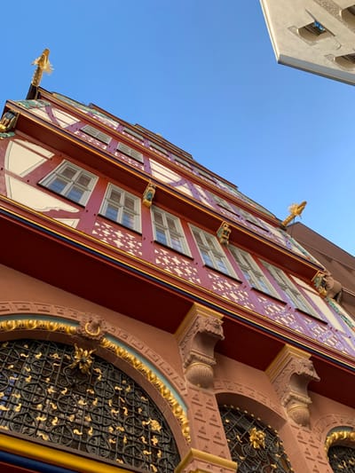 The image shows the facade of a building located at Römerberg, in the city of Frankfurt am Main, Germany. The building has a red, white, and gold facade with intricate details and multiple windows. There are gold-painted statues on the facade and some of the windows are adorned with ornate wrought iron grilles. The image is taken from a low angle looking upwards toward the sky. The sky is blue and clear. The top edge of another building is visible at the top of the image, to the left of the sky. The image appears to be taken during the daytime. There are no people visible in the image.