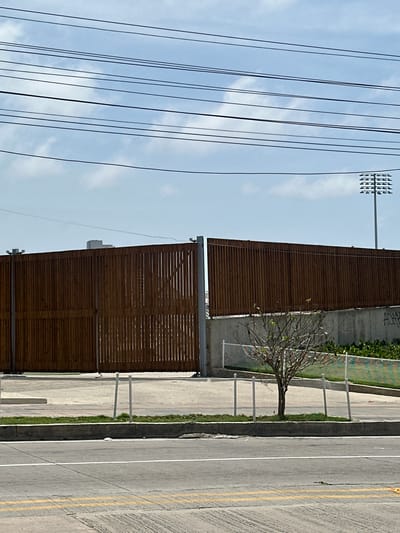 The image shows a large wooden gate, with vertical planks, that is partially open. The gate is situated in front of a concrete wall, behind which is a lush green area with a small, leafless tree in the foreground. To the right of the gate, a stadium light on a tall pole is visible against a cloudy blue sky. Overhead, several power lines stretch across the frame. In the foreground, a street with yellow and white markings is visible, bordering a sidewalk with a low white fence. The overall scene suggests an urban environment, possibly near a sports facility. The location is identified as Perímetro Urbano Barranquilla, Colombia.