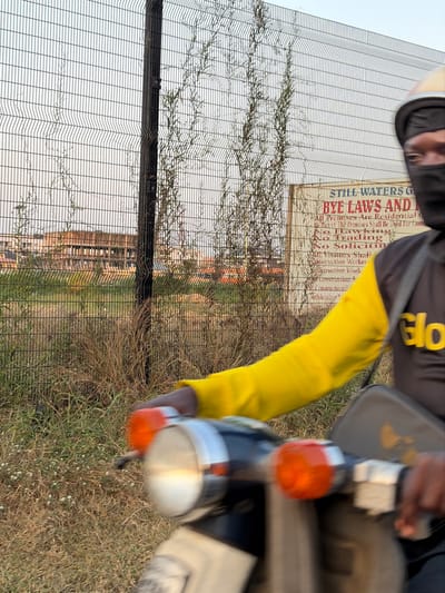 A blurred image, rotated 90 degrees, shows a person in yellow and black clothing near a wire fence with growing vines. A sign on the fence reads "STILL WATERS AND". Buildings can be seen through the fence. The front of a motorbike with blurred lights and a windshield is in the foreground, obscuring some of the scene. The background includes grass.