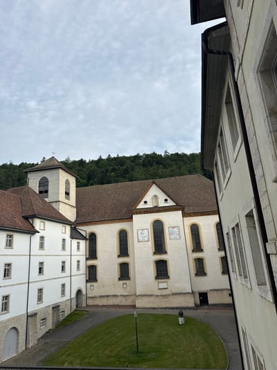 The image displays a view of the Bellelay Abbey in Bellelay, Switzerland, under a cloudy sky. The foreground features a segment of a building with a light-colored facade and multiple windows. To the left, a white building with red-tiled roofs and a bell tower is visible. The bell tower is square with a pointed roof and a weather vane. Adjacent to it is a larger church building with a brown-tiled roof and arched windows. The church facade is cream-colored and adorned with two sundials. A well-maintained green lawn with a paved walkway is situated in the courtyard between the buildings. In the background, a dense forest covers a hillside. The overall scene depicts the historic architecture of the abbey complex.
