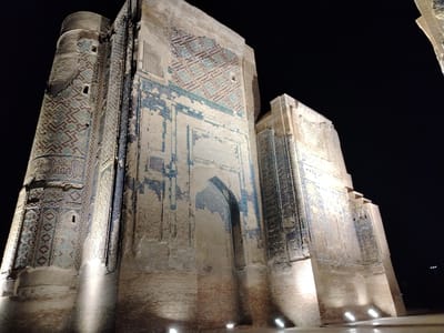 The photograph captures an exterior view of the Ak-Saray Palace complex in Shahrisabz, Uzbekistan, at night. The main subject is a section of the palace's grand, ancient architecture, characterized by its massive brick construction adorned with intricate blue and turquoise geometric tilework. The lighting from ground-level spotlights illuminates the facade, casting dramatic shadows and highlighting the aged texture of the bricks and the detailed patterns of the tiles. The dark night sky serves as a stark background, emphasizing the illuminated structures. The perspective is low-angle, looking up at the imposing scale of the building. No people or discernible actions are visible, focusing solely on the architectural grandeur of the historical site.