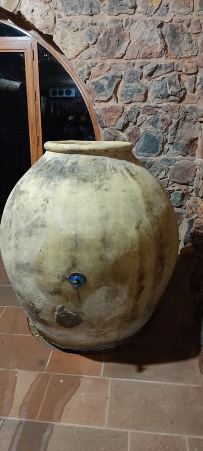 An aged, light-colored, large clay amphora with a tap attached to its side is the main subject of this image, positioned on a terracotta tiled floor. The amphora exhibits a weathered patina with darker discoloration and visible texture. Engraved on its upper body is a date, "1976", flanked by decorative lines. The background features a rustic stone wall constructed from irregular, multi-colored stones with visible mortar. To the left of the amphora, a wooden framed glass door with an arched top is visible, reflecting interior elements such as what appear to be cooling units and possibly people dressed in casual attire. The scene is indoors, with the lighting casting shadows that suggest a dimly lit environment. This setting is identified as being located in Taperakan, Armenia.