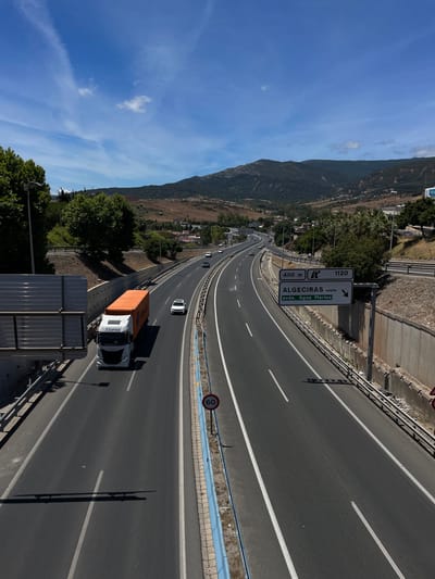 This is a photograph of a highway in Algeciras, Spain, taken during the day. The highway curves through a landscape with a mountain range on the left and trees and buildings on the right. The sky is blue with some scattered clouds.

Several vehicles are visible on the highway, including a white truck with an orange container and the logo "Alonso" on its side, a white car, and several other cars in the distance. A road sign indicates "ALGECIRAS oeste" and "avda. Agua Marina" along with distances of "400 m" and "1120". There is also a speed limit sign showing "60".

The image appears to be taken from an elevated position, possibly an overpass or a higher point of the highway, looking down and along the road. The sunlight casts shadows on the road, suggesting it is mid-morning or afternoon. The overall scene depicts a typical highway commute in a Mediterranean setting.
