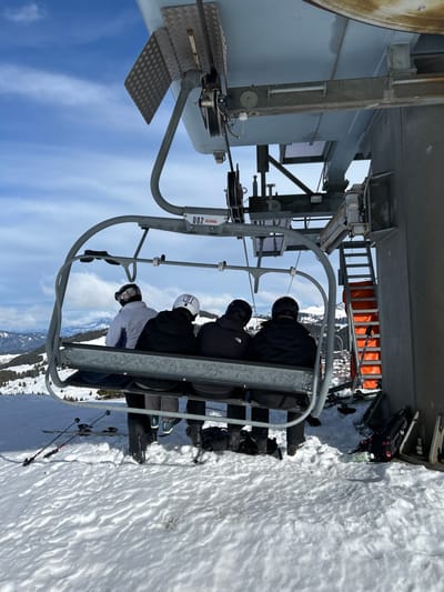 Four individuals are depicted from behind, seated on a grey metal ski lift chair at a mountain ski resort in Villard-sur-Doron, France. The chair, marked with "002" and "SKIRAIL", is positioned at a pylon structure that forms part of the lift mechanism, suggesting a loading or unloading station. The pylon, on the right, is a large grey structure with exposed metal platforms, machinery, and an attached metal ladder featuring orange safety padding.
All four individuals wear ski helmets and jackets; the leftmost person has a white helmet and jacket, while the other three wear black helmets and dark-colored jackets. Their legs extend down, with ski boots visible, though skis or snowboards are not attached to their feet.
The ground is covered in undisturbed snow. To the left of the chair, a pair of skis with poles lies flat on the snow. To the right, a snowboard and a black backpack rest on the snow, near the pylon, alongside another pair of skis with poles leaning against the pylon and orange ladder.
In the background, under a blue sky with scattered white clouds, snow-covered mountains dotted with evergreen trees extend into the distance. Faint structures, possibly part of a village or resort, are visible in the far background on the right.