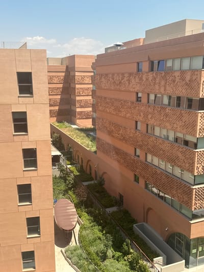 This image shows a modern architectural complex in Thessaloniki, Greece. The buildings are constructed with a warm, terracotta-colored material and feature a distinctive patterned brickwork facade. The complex includes multiple stories with numerous windows, some of which are open.

Below the buildings, a lush green courtyard area is visible. This courtyard features neatly manicured vegetation, including trees and bushes, and a long, oval-shaped, dark-colored outdoor table. The courtyard is bordered by a fence and includes paved walkways.

In the background, other buildings of similar architectural style can be seen, some with rooftop amenities. The sky is clear with a few scattered clouds, suggesting a bright, sunny day. The perspective of the image is from a slightly elevated viewpoint, looking down into the courtyard and at the facades of the buildings.

The location is identified as the Alexander Innovation Zone (AIX) in Thessaloniki, Greece. AIX is a hub for innovation and technology, aiming to foster collaboration and development in the city. The architecture reflects a contemporary design that integrates green spaces with functional buildings.