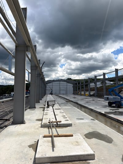 The image depicts an outdoor construction site in Stróża, Poland, characterized by unfinished steel structures and concrete foundations under a dramatic, cloudy sky. In the foreground, large gray steel beams are erected, supported by concrete pillars. The ground is a mix of concrete slabs and gravel, with some areas featuring puddles of water, suggesting recent rain. Several concrete elements are laid out, some with metal rebar protruding. A long, shallow channel of water runs alongside the constructed pathway. In the midground, a white building with a large door is visible, along with construction equipment including a blue boom lift (Genie brand) and cranes. The background shows rolling green hills and trees. There are no people clearly visible in the frame, but the presence of construction machinery indicates ongoing work.