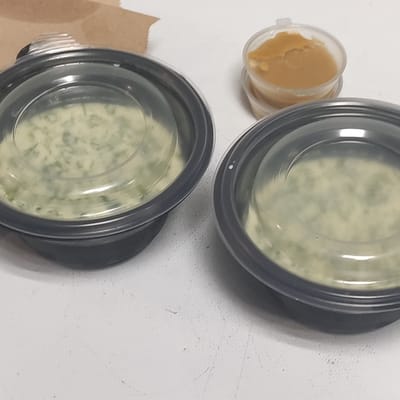 Two black plastic containers with clear plastic lids sit on a white surface. Each container is filled with a light green, creamy substance with small dark green flecks. To the right of the containers is a small, clear plastic cup filled with a light brown, semi-solid substance. A brown paper bag is visible in the upper left corner of the image. The image appears to be a close-up shot of food items.