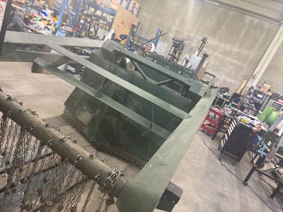 The image displays an indoor industrial workshop or fabrication facility. The primary subject is a large, angular piece of machinery, olive-green in color, composed of heavy-gauge metal panels. This structure features a robust central body with sloped sides and a base incorporating perforated metal plating in a honeycomb pattern. In the immediate foreground, a horizontal metal bar is affixed with numerous short, linked chains, each terminating in a small, round metal disk, suggesting a flail or shredding mechanism. Multiple large springs are visible on top sections of the green machinery, implying a suspension or vibratory function. A second, similar green metal structure is partially visible further into the background.

One individual is partially visible in the mid-ground, positioned behind the main green machinery. Only the head and upper torso are discernible; the individual is wearing a white hooded garment. Their face is turned towards the viewer or camera.

The environment consists of bare concrete walls and a concrete floor. The background is an active workshop space, cluttered with various tools, equipment, and materials. Notable items include multi-tiered metal shelving units filled with miscellaneous objects, multiple workbenches holding tools and components, rolling tool carts (one red, one black with drawers), and a large spool of green filament on a stand. A blue cable runs across the concrete floor. The precise location within the city of Logatec, Slovenia, cannot be determined from the visual information provided.