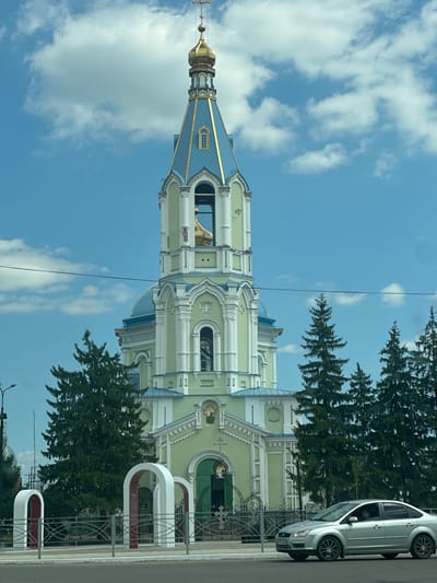 The image shows the Ascension Cathedral in Rasskazovo, Russia, on a sunny day with scattered clouds. The cathedral features a tall, light green bell tower with white trim and a blue roof topped with a golden dome and cross. Several tall evergreen trees stand in front of the cathedral. In the foreground, a silver sedan is parked on the side of the road, and a metal fence encloses a pathway leading to the cathedral entrance, which is marked by three white arched structures. A power line stretches across the sky above the scene. No people are visible.