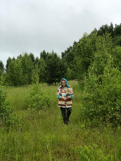 A person wearing a blue headscarf and a colorful patterned jacket walks through a grassy field with trees in the background. The person is holding a small bouquet of flowers. The sky is overcast. This image was taken in the city of Zhukovo, Russia.