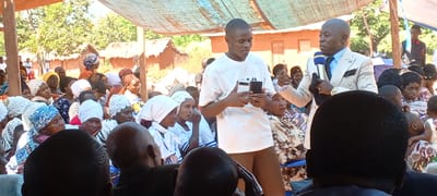 The image shows a gathering of people in an outdoor setting in Kalemie, Democratic Republic of the Congo.
In the foreground, several people sit facing forward, with the backs of their heads visible. A young man in a white T-shirt and brown pants stands holding a smartphone. Beside him, an older man in a light-colored suit with a blue tie holds a microphone and speaks.
In the background, numerous individuals are seated, including many women wearing head coverings. Some of the women wear white outfits and head coverings. Beyond the crowd, there are buildings and trees under a partly cloudy sky. An awning provides shade for those gathered.