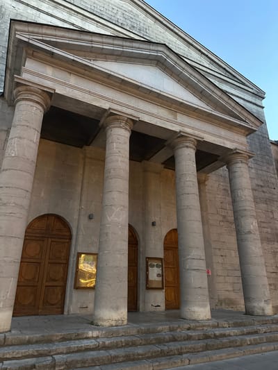 The image shows the facade of a stone building with classical architectural elements. There are several large, fluted stone columns supporting a triangular pediment. The entrance to the building appears to be through large wooden double doors, with at least two sets visible. There are also display cases mounted on the wall between the doors. The building is constructed from light-colored stone blocks, and the overall impression is one of grandeur and age. The sky above is a clear blue, suggesting daylight.

The building depicted is the Protestant temple in Anduze, France.