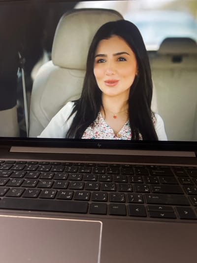 The image displays an HP laptop, dark gray in color, with its screen showing a video. The laptop's keyboard, featuring both Arabic and Latin characters, and a section of its silver-gray palm rest are visible in the lower portion of the frame. The video depicts a young adult female with long dark hair, a light complexion, and noticeable makeup including eyeliner and lipstick. She is wearing a white collared shirt layered over a floral patterned top and a thin gold chain necklace with a small red heart pendant. She is seated within what appears to be a vehicle interior, as indicated by light-colored (cream or beige) car seats visible behind her. Her expression is a slight smile, and her gaze is directed slightly off-camera to her left. The environment outside the vehicle, visible through a window, is blurred. The specific location of Nuseirat, Palestinian Territory, cannot be determined from the visual information provided in this image.