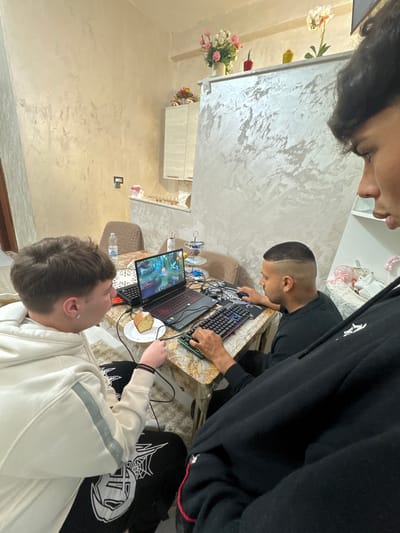 Three individuals are gathered around a table indoors. One person is seated wearing a white sweatshirt, another in a black hoodie, and the third is partially visible in the upper left corner. The person in the black hoodie is using a computer with a keyboard and mouse. A laptop displaying a game is open on the table, beside a plate of sliced cake. A water bottle is also visible on the table. The room features textured beige walls, floral decorations, and a visible section of kitchen cabinets. The location is Stornara, Italy.