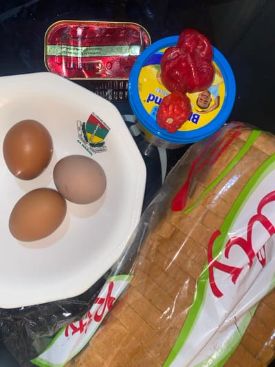 An overhead view in Tudun Nupawa, Nigeria, displays a collection of food items arranged on a dark, reflective surface. A white octagonal bowl, featuring a logo inscribed with "ALL LIVES ARE PRECIOUS" and "44 NARHK KADUNA," contains three brown-shelled eggs. To the upper left, a red and gold rectangular can of "OGBONGE" brand sardines in olive oil is visible. Adjacent to the right, a blue-lidded tub of "Blue Band" margarine or spread, depicting a child's face on its lid, is topped with two red peppers, one large and one smaller, possibly scotch bonnets. A clear plastic bag containing a sliced loaf of white bread with a red and green label partially visible occupies the lower right portion of the frame. No individuals are present, nor are any actions or interactions depicted.